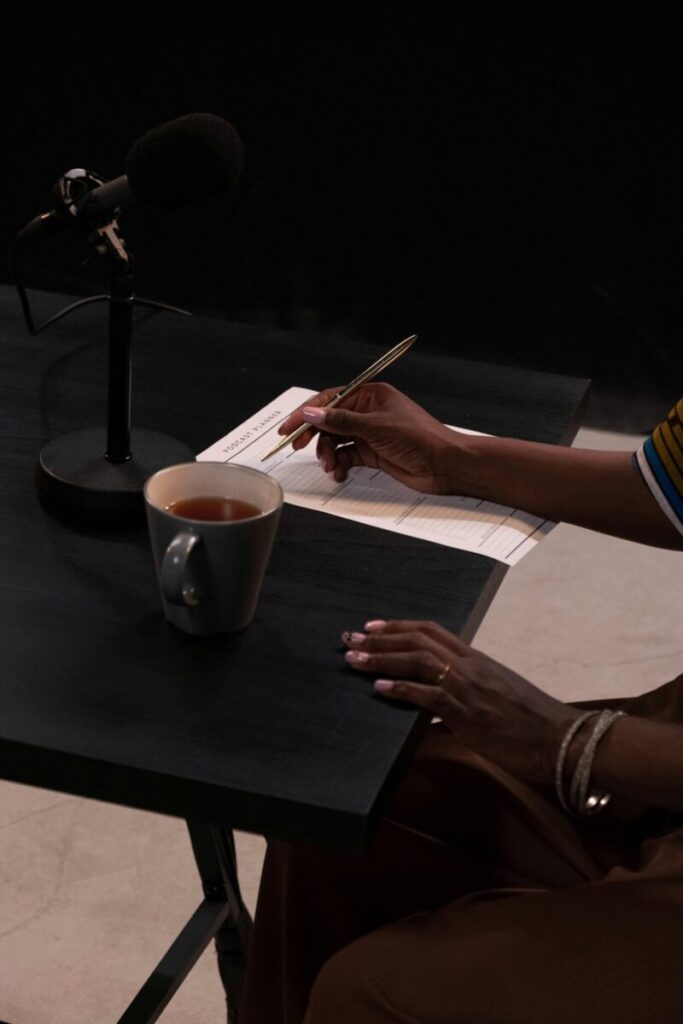 A person recording a podcast with a microphone, cup of tea, and notes on a table.